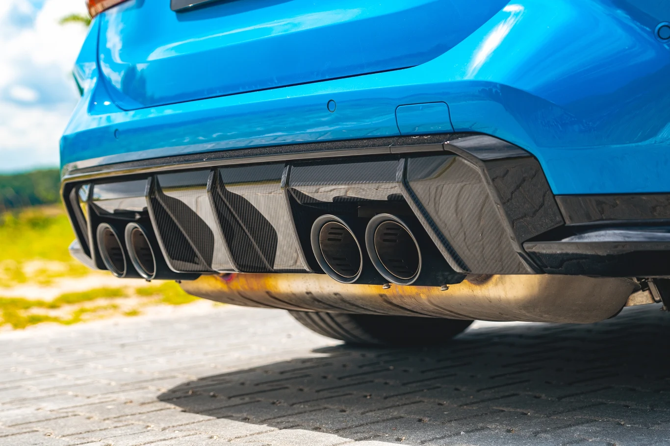 Dolna część sportowego samochodu w niebieskim kolorze z widocznymi czterema końcówkami układu wydechowego i czarnym dyfuzorem, auto stoi na brukowanym podłożu.