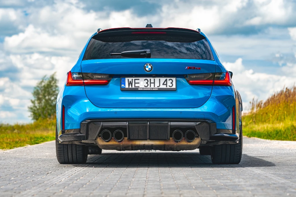 Błękitny samochód BMW M3 Touring widoczny od tyłu, auto stoi na brukowanej drodze, w tle widać trawę i niebo z chmurami, charakterystyczne tylne lampy LED oraz sportowy układ wydechowy.
