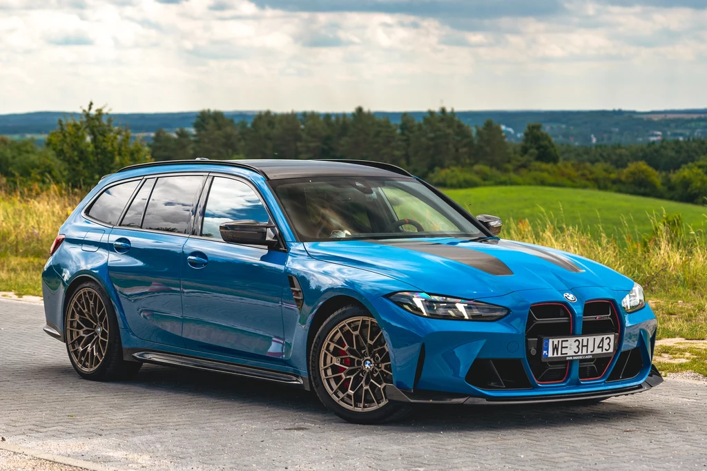 Niebieskie sportowe kombi BMW M3 Touring zaparkowane na tle zielonego krajobrazu i rozległych pól, nowoczesny design auta podkreślają czarne felgi oraz kontrastujące detale.