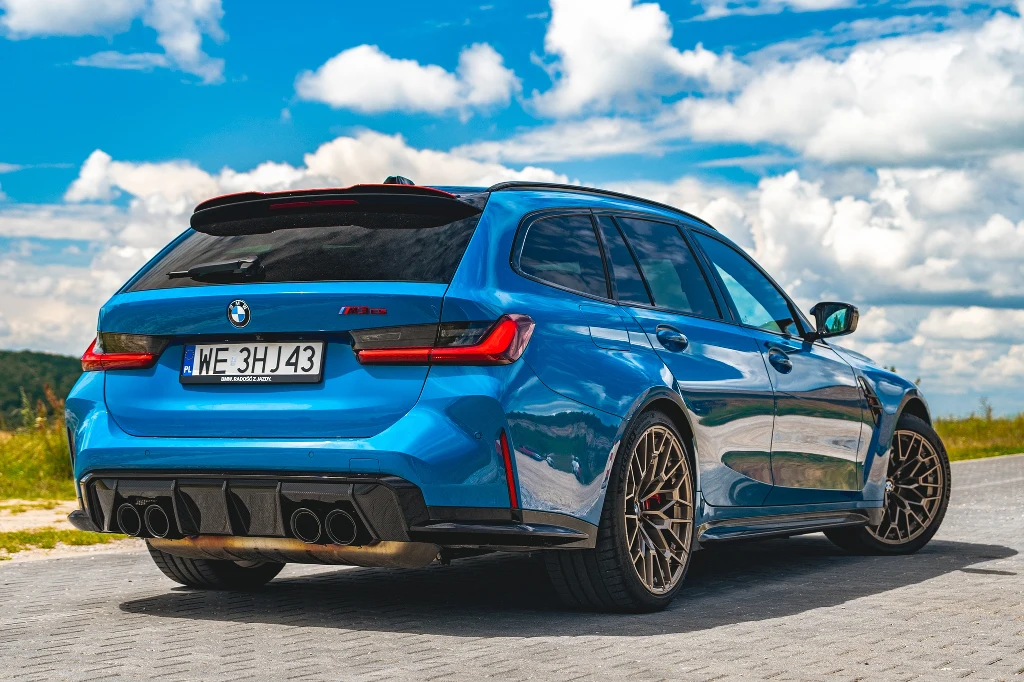 Niebieskie kombi marki BMW M3 zaparkowane na kostce brukowej, w tle letni krajobraz z niebem wypełnionym chmurami, samochód wyróżnia się dynamiczną sylwetką i sportowymi felgami.