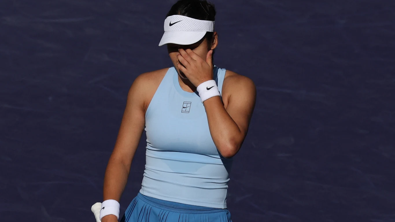 Tenisistka w jasnoniebieskim stroju sportowym zasłania twarz dłonią, trzymając rakietę tenisową w lewej ręce. W tle widoczna ciemna powierzchnia kortu.