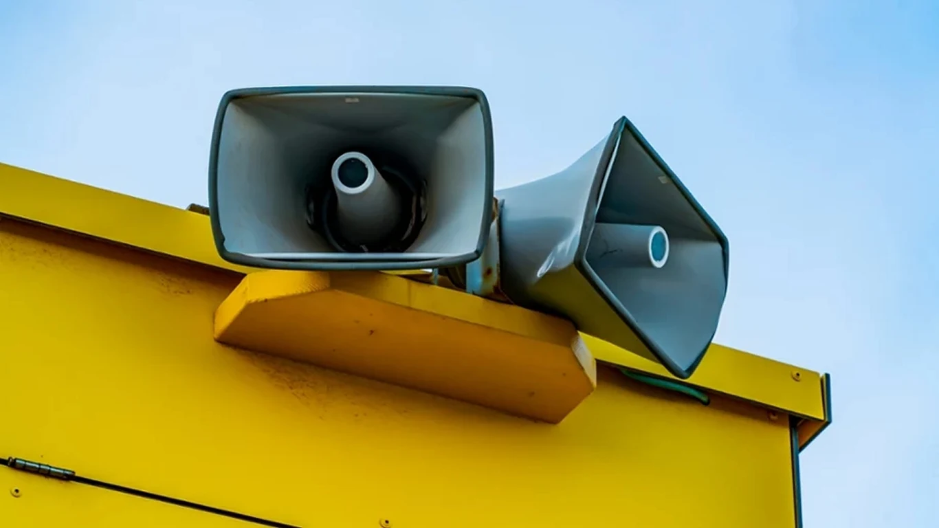 Co zrobić, gdy syreny zawyją w Twoim mieście i jak reagować na alerty RCB? Dwa szare głośniki przemysłowe zamontowane na żółtym metalowym dachu budynku, skierowane w różnych kierunkach, na tle błękitnego nieba.