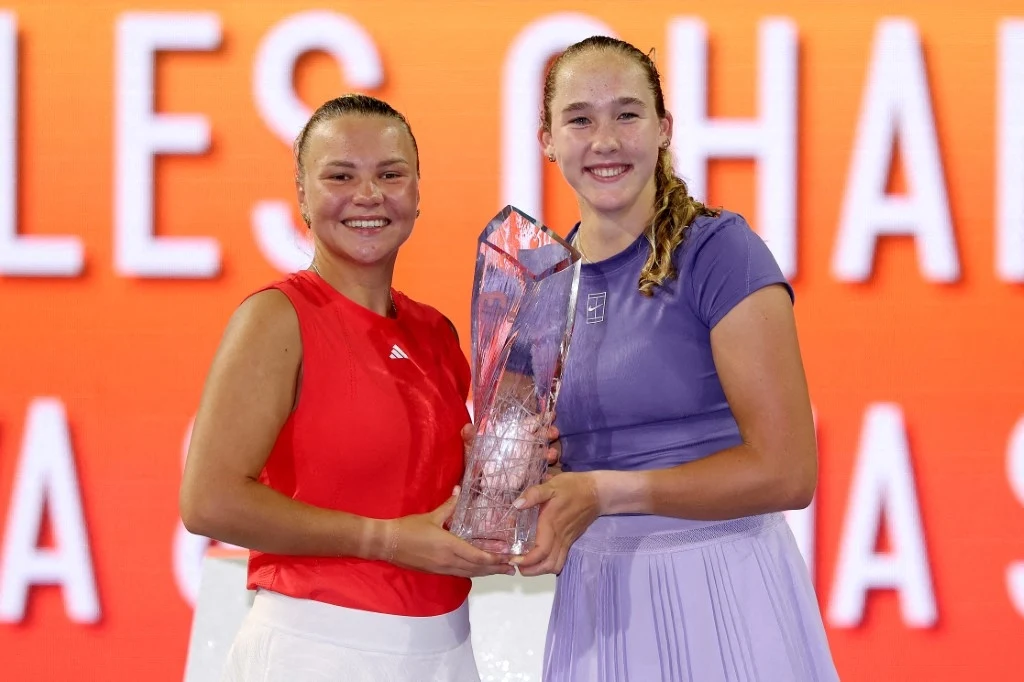 Diana Sznajder i Mirra Andriejewa Dwie młode kobiety stoją obok siebie i trzymają wspólnie kryształowy puchar, obie uśmiechnięte, ubrane w sportowe stroje na tle pomarańczowej tablicy z białym napisem.