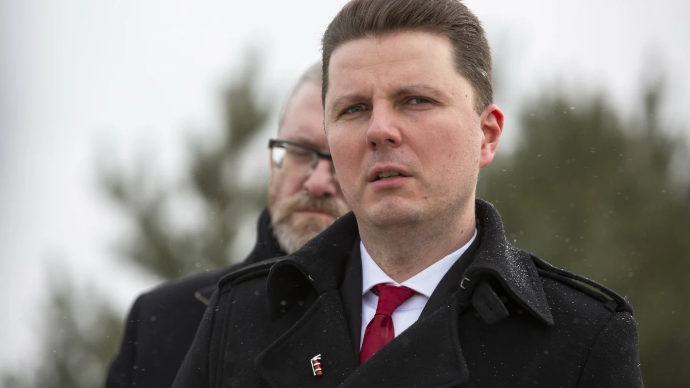 Mężczyzna w ciemnym płaszczu i czerwonym krawacie na pierwszym planie, w tle kolejne osoby, rozmyte drzewa oraz zimowa sceneria.