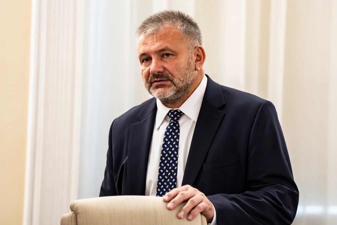 Waldemar Żurek Mężczyzna w średnim wieku w garniturze i krawacie stojący za jasnym krzesłem, w tle jasne zasłony.