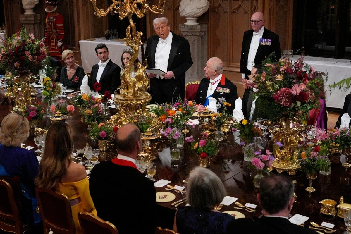 Uroczysta kolacja w eleganckiej sali, przy bogato ozdobionym stole siedzą zaproszeni goście w wieczorowych strojach, a mężczyzna w smokingu wygłasza przemówienie na stojąco.