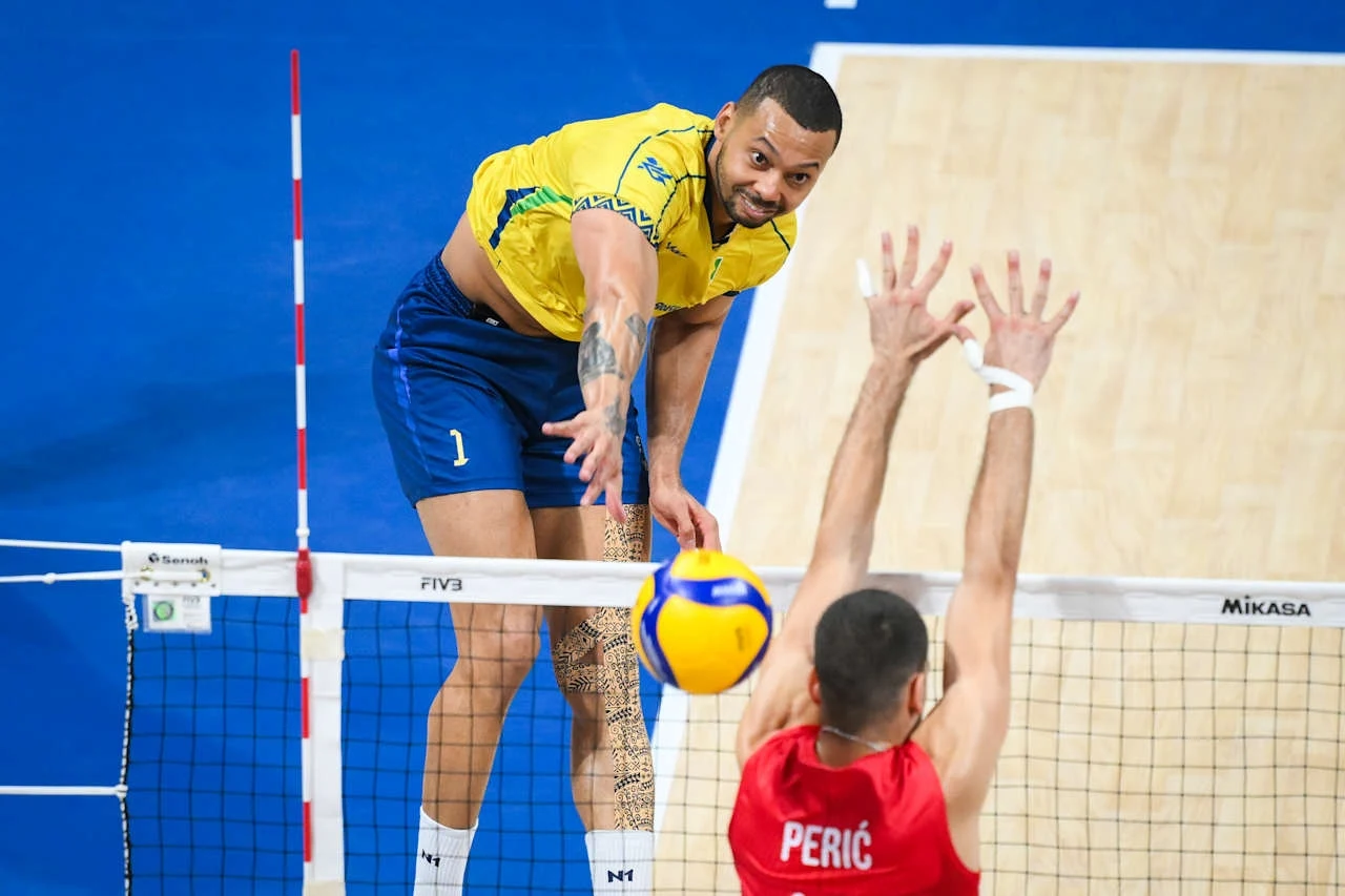 Alan Souza Zawodnik w żółto-niebieskim stroju atakuje piłkę ponad siatką podczas meczu siatkówki, a naprzeciwko zawodnik w czerwonym stroju z nazwiskiem Perić na plecach próbuje zablokować atak.