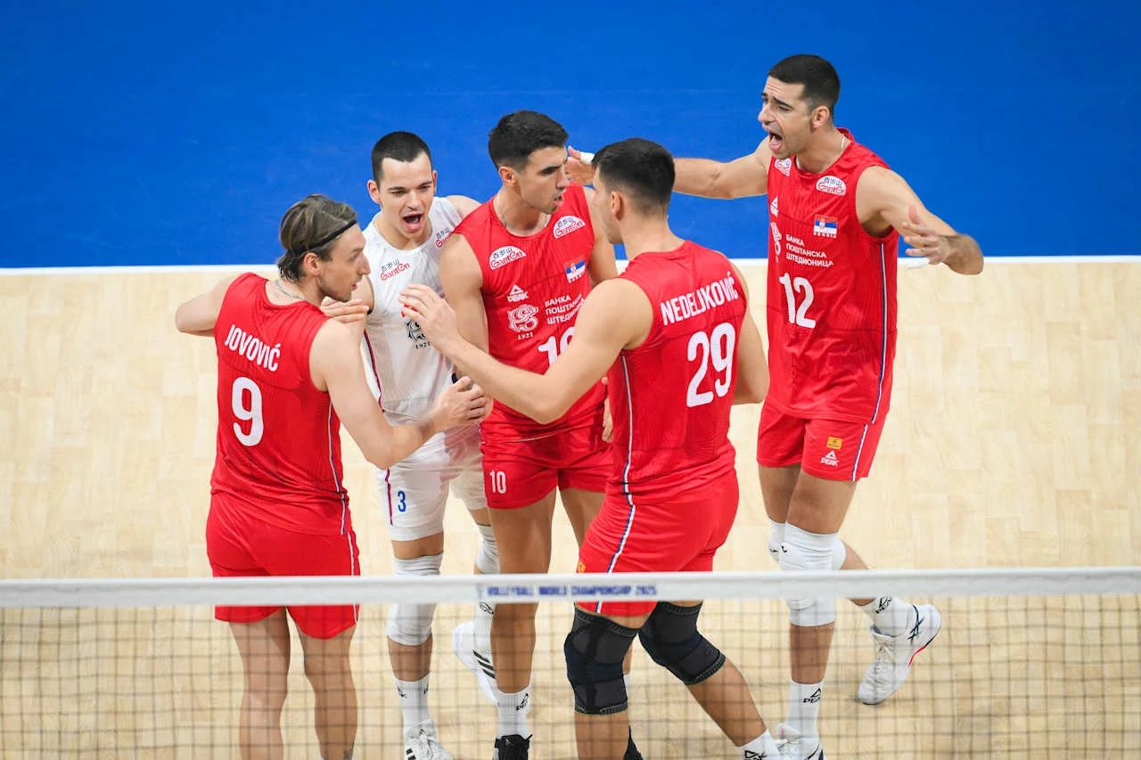 Mecz Brazylia - Serbia zakończył się sensacyjnie Grupa siatkarzy w czerwonych i białych strojach celebruje udaną akcję na boisku, wszyscy są widocznie zaangażowani i wyrażają radość gestami i mimiką.