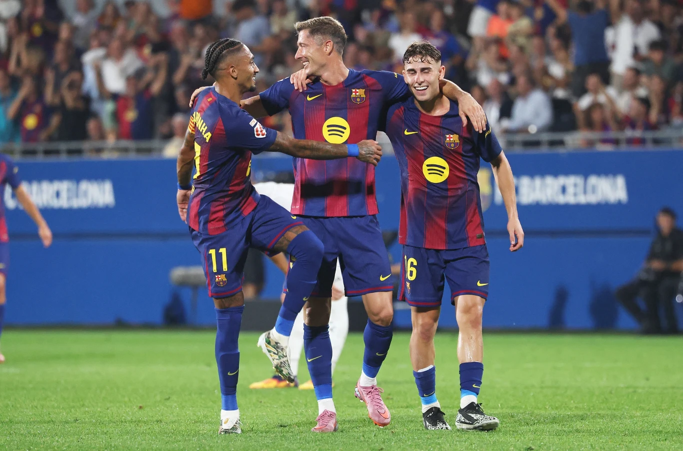 Trzech piłkarzy FC Barcelony świętujących bramkę na boisku, uśmiechnięci, z ramionami na swoich barkach, ubrane w klubowe stroje w granatowo-bordowe pasy, w tle kibice na trybunach.