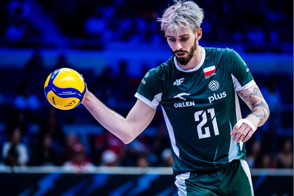 Siatkarz w zielonym stroju z numerem 21 trzyma piłkę do siatkówki, skoncentrowany przed serwisem, na jego koszulce widoczne logotypy sponsorów oraz flaga Polski, rozgrywka odbywa się w hali sportowej z publicznością w tle.