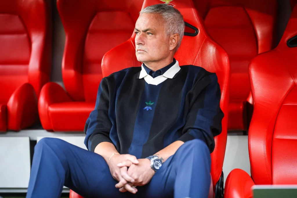 Jose Mourinho Mężczyzna z siwymi włosami w marynarce i ciemnych spodniach siedzi na czerwonym fotelu stadionowym, patrząc skupionym wzrokiem w bok; za nim rząd identycznych, pustych foteli.