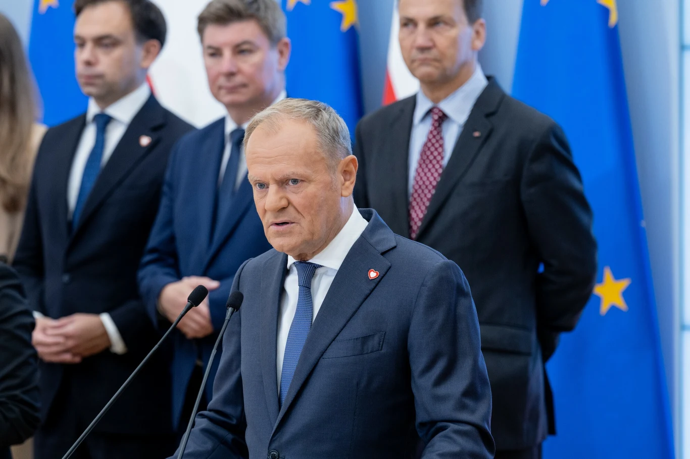 Rząd bierze się za uszczelnianie podatków, nie trzymając się przy tym obietnicy 6-miesięcznego vacatio legis dla zmian podatkowych Grupa polityków w garniturach stojących na tle flag europejskich i narodowych, mężczyzna przy mównicy wygłasza przemówienie, pozostali słuchają.