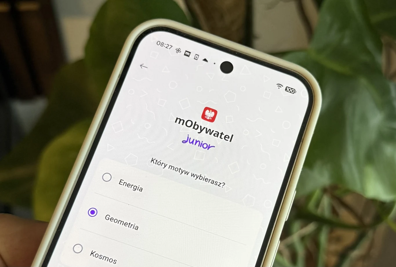 mObywatel Junior dostępny. Cyfrowa legitymacja szkolna na smartfonie. Ekran smartfona pokazujący aplikację mObywatel Junior z widoczną listą tematów do wyboru, w tym zaznaczoną opcją 'Geometria', w tle rozmazane liście rośliny.