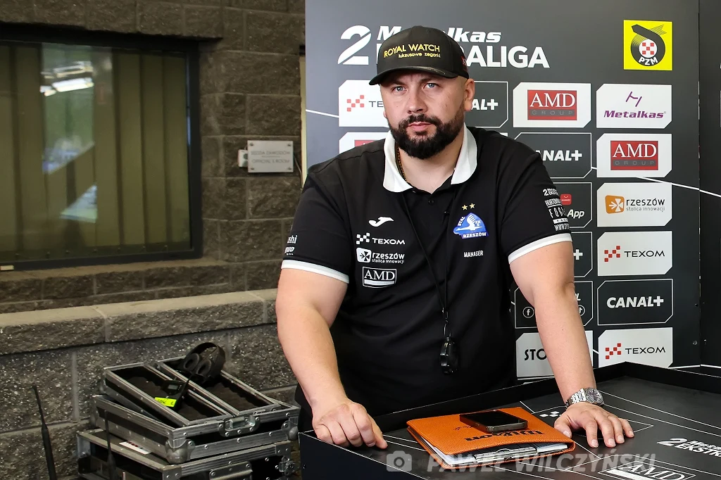 Paweł Piskorz, menadżer Texom Stali Rzeszów. Mężczyzna w czarnej koszulce i czapce z daszkiem stoi przy stole z laptopem, w tle ścianka z logotypami sponsorów oraz sprzęt techniczny. Na odzieży widoczne emblematy klubowe i sponsorskie, w tle wyraźne akcenty związane z wydarzeniem sportowym.
