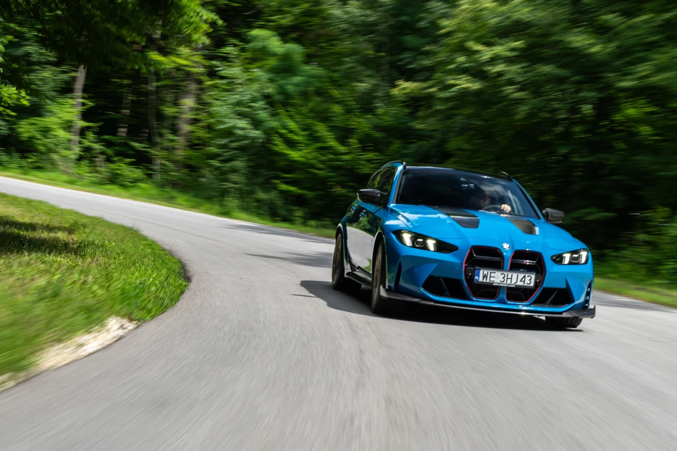 Niebieski sportowy samochód BMW jedzie z dużą prędkością po krętej, asfaltowej drodze otoczonej bujną zielenią i drzewami, podkreślając dynamikę jazdy oraz techniczne możliwości pojazdu.