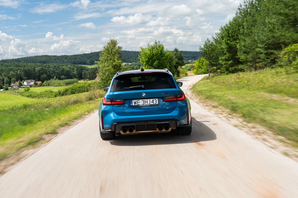 Niebieskie sportowe kombi BMW jedzie wąską, asfaltową drogą pośród zieleni, z lasem po jednej stronie i otwartym terenem rolniczym po drugiej, przy słonecznej pogodzie na tle lekkiego, pochmurnego nieba.
