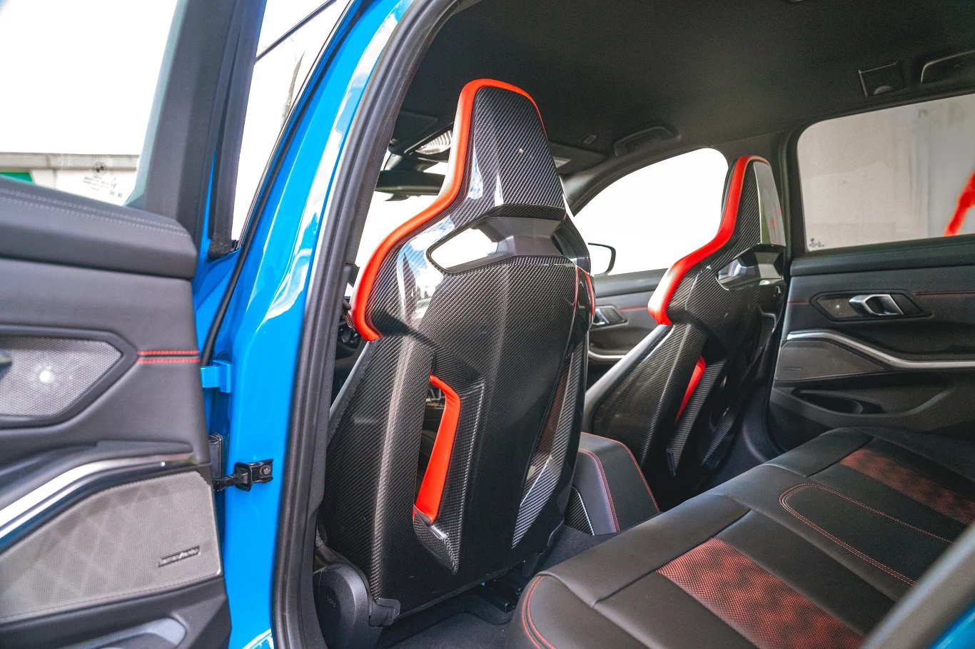 Wnętrze nowoczesnego samochodu z widokiem na tylne siedzenia, pokrycia foteli wykonane z czarnego materiału z czerwonymi akcentami oraz plecy przednich foteli z widoczną karbonową strukturą i detalami w kolorze czerwonym.