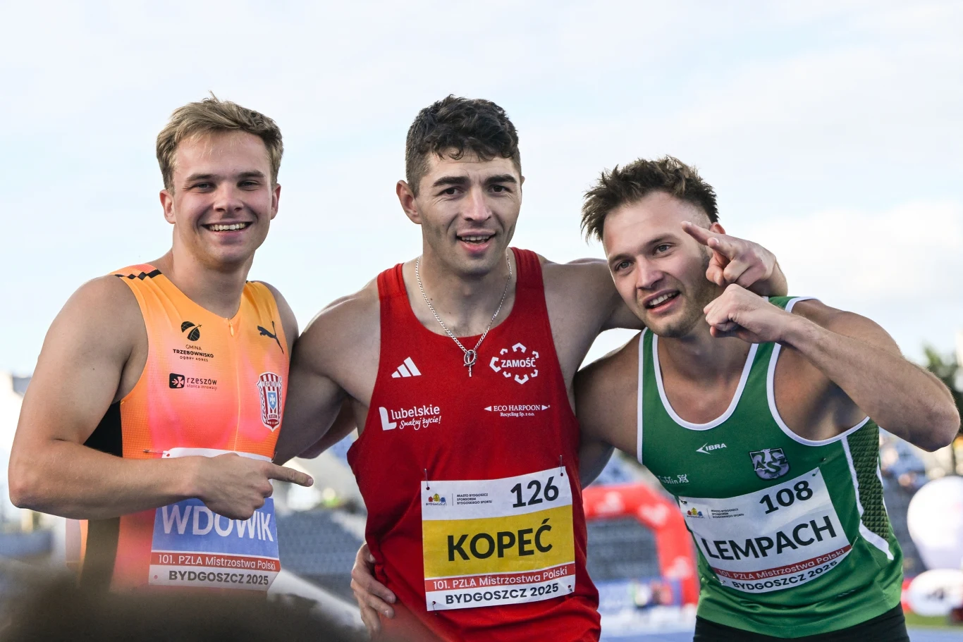 Trzech młodych sportowców pozuje razem na stadionie, ubrani w stroje startowe w różnych kolorach, jeden z nich stoi w środku i trzyma dwóch pozostałych za ramiona, wszyscy uśmiechają się do aparatu.