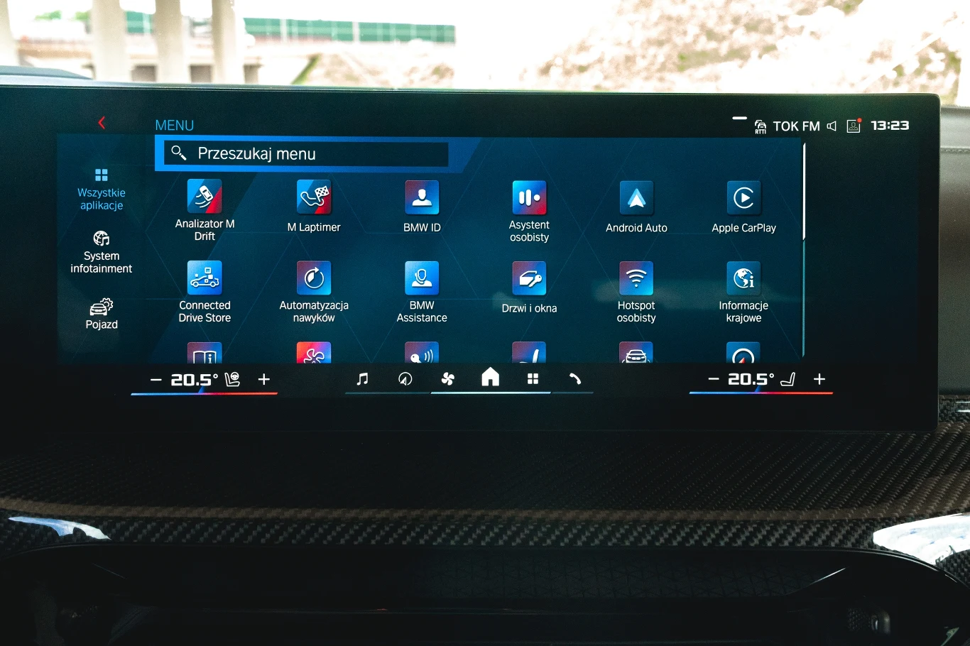 Dotykowy ekran systemu multimedialnego BMW z wyświetlonym menu aplikacji, widoczne ikony funkcji takich jak nawigacja, asystent osobisty, Apple CarPlay, Android Auto oraz ustawienia klimatyzacji ze wskazaniem temperatury dla dwóch stref.