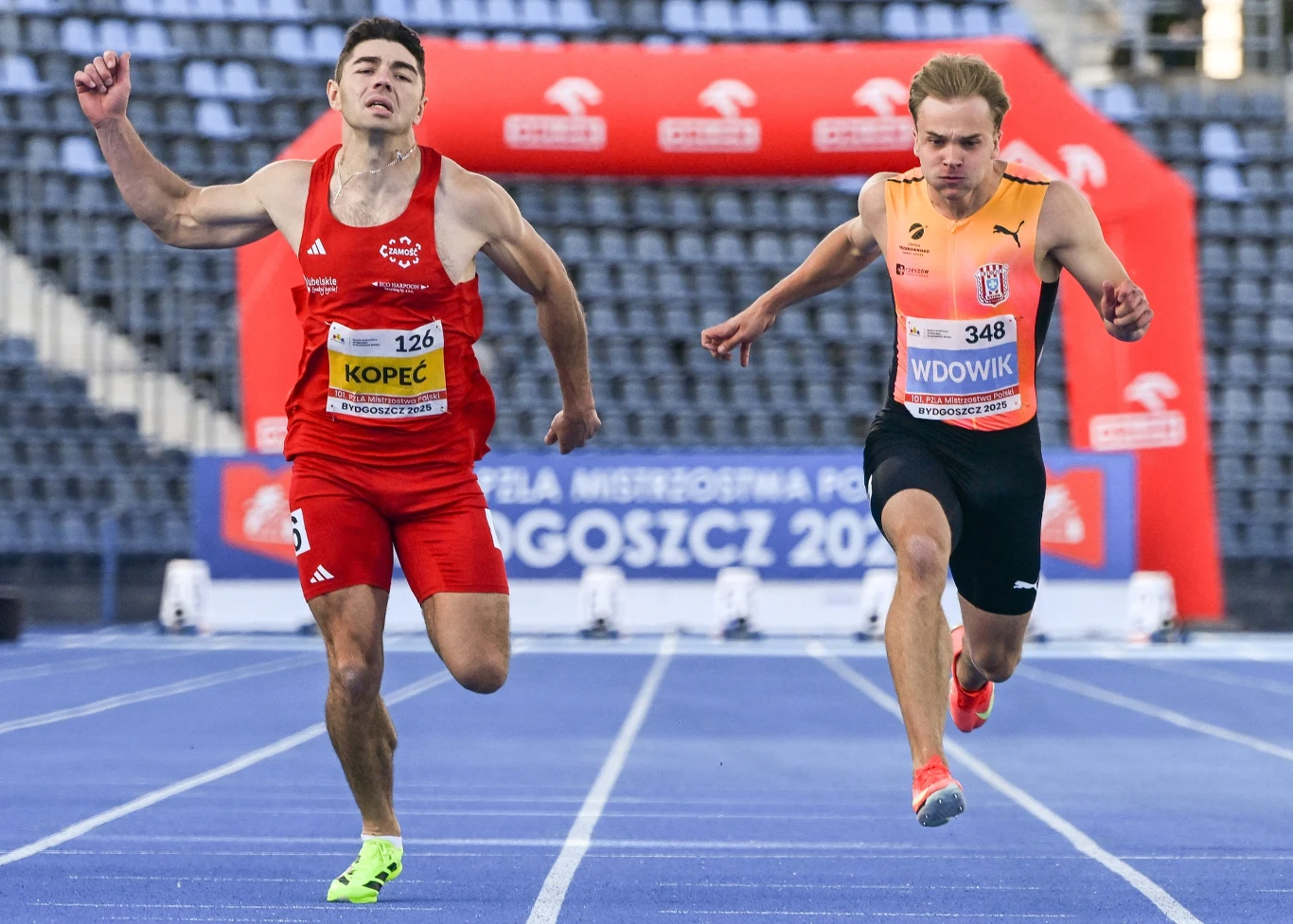 Dwóch lekkoatletów ścigających się na bieżni tuż przed metą, widoczne emocje na ich twarzach, intensywny wysiłek fizyczny, trybuny stadionu oraz brama z napisem 'Bydgoszcz 2021' w tle.