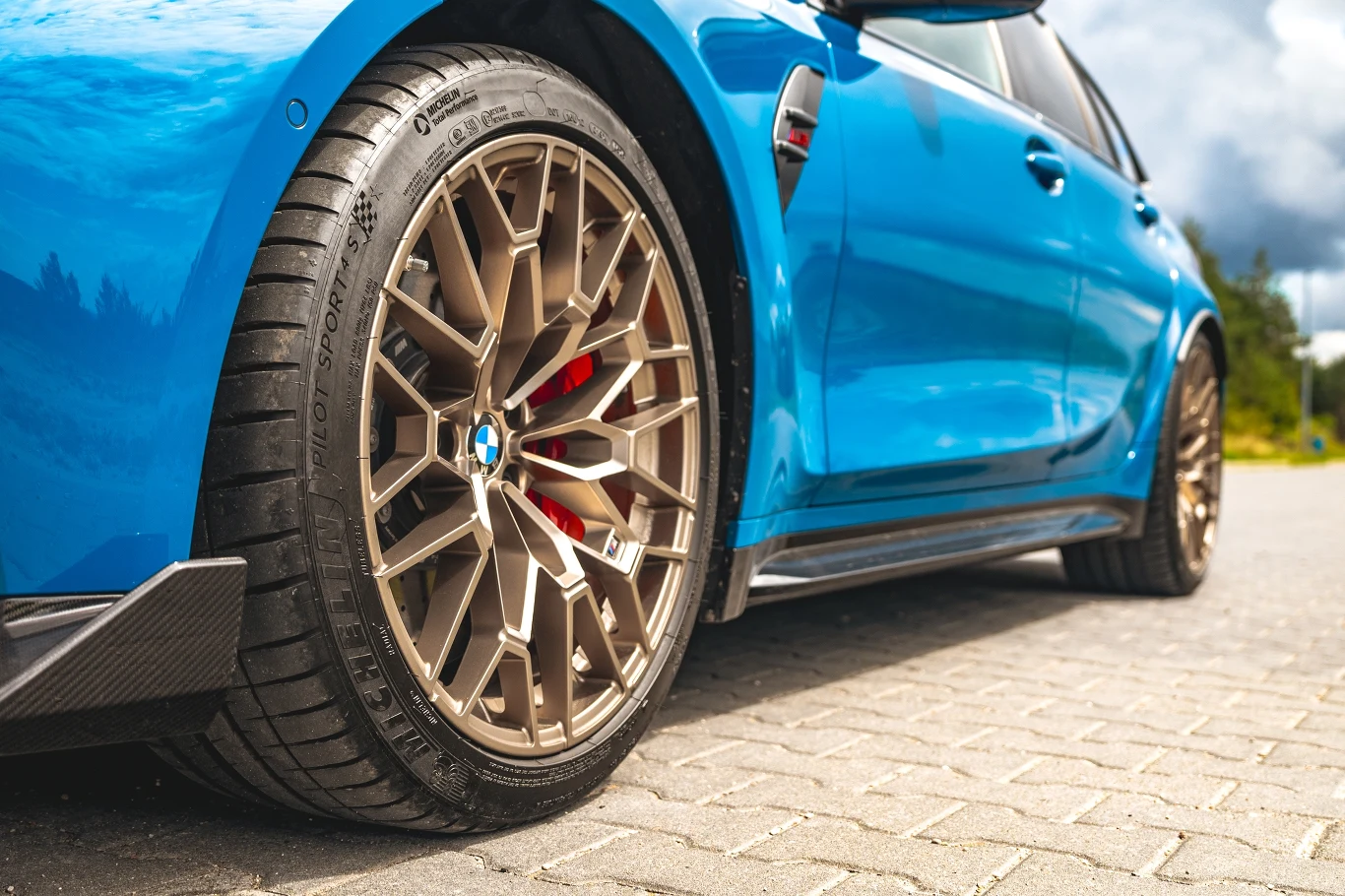 Niebieski samochód sportowy BMW z wyeksponowanymi złotymi felgami i dużymi wentylowanymi hamulcami zaparkowany na kostce brukowej.