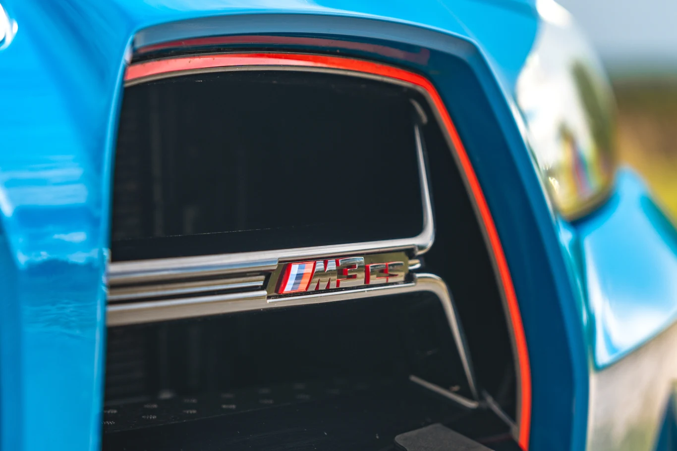 Detal z przodu samochodu BMW M3 w kolorze niebieskim, z wyraźnie widocznym logo serii M3 na kratce wlotu powietrza oraz akcentami kolorystycznymi w odcieniu czerwieni.