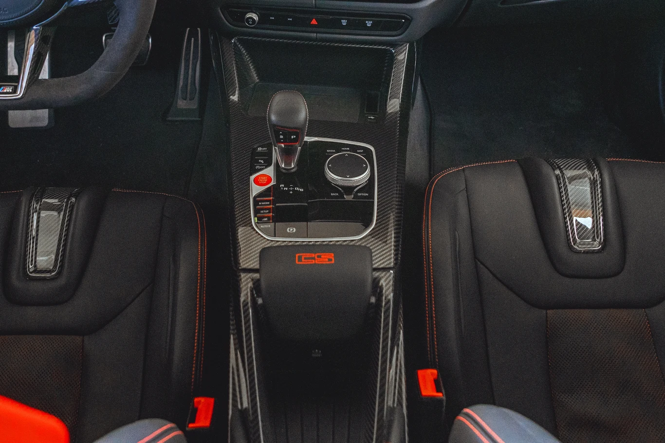 Kokpit sportowego samochodu z karbonowym wykończeniem tunelu środkowego, widoczna dźwignia zmiany biegów, pokrętło sterowania multimediami oraz dwa kubełkowe fotele z czerwonymi przeszyciami.