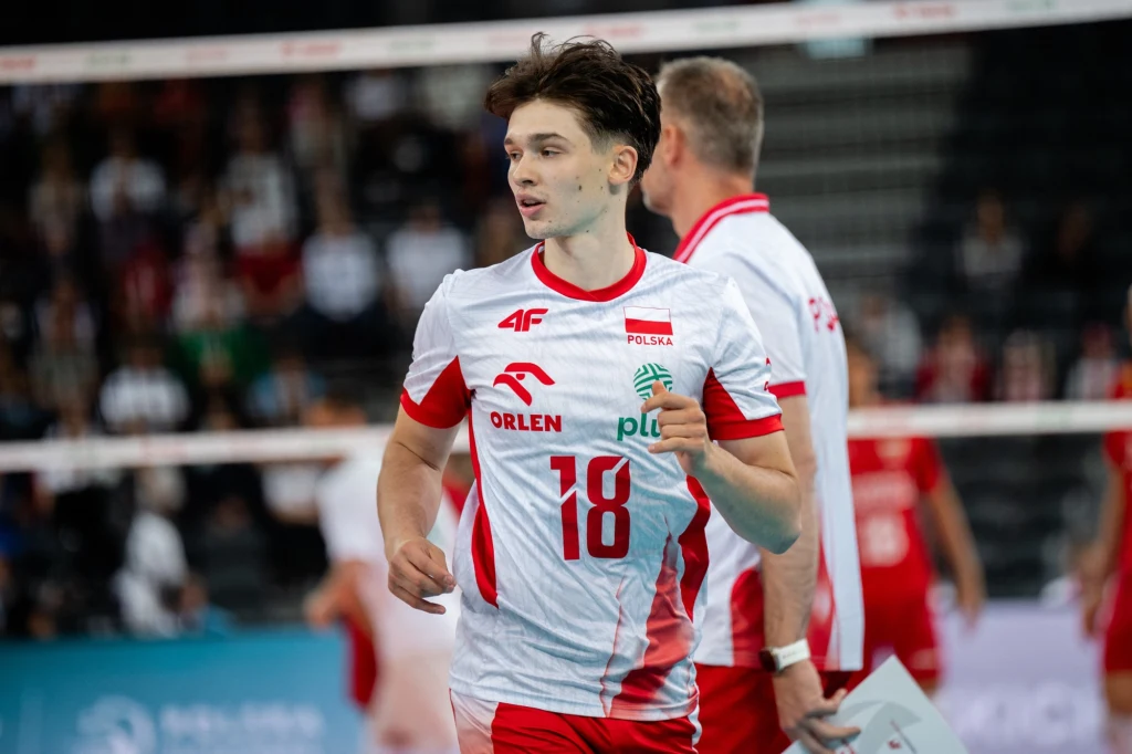 Maksymilian Granieczny Polski siatkarz w stroju reprezentacji Polski na boisku siatkarskim, w tle widoczni inni zawodnicy podczas meczu.