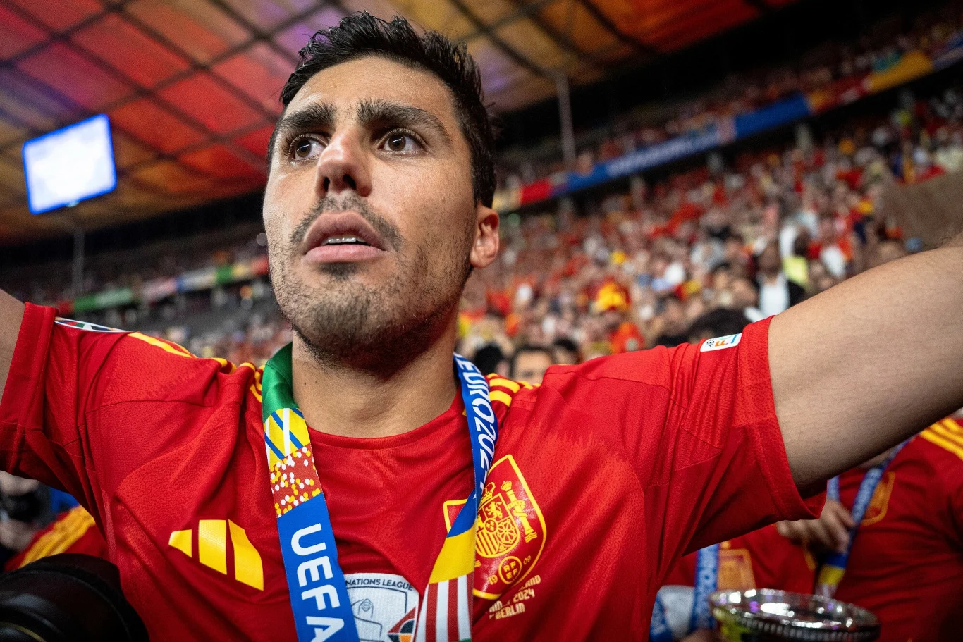 Rodri Piłkarz w czerwonej koszulce reprezentacji Hiszpanii z medalem UEFA na szyi, stojący na stadionie pełnym kibiców, unosi ręce w geście triumfu. W tle dużo osób ubranych w czerwone barwy, atmosfera zwycięstwa.