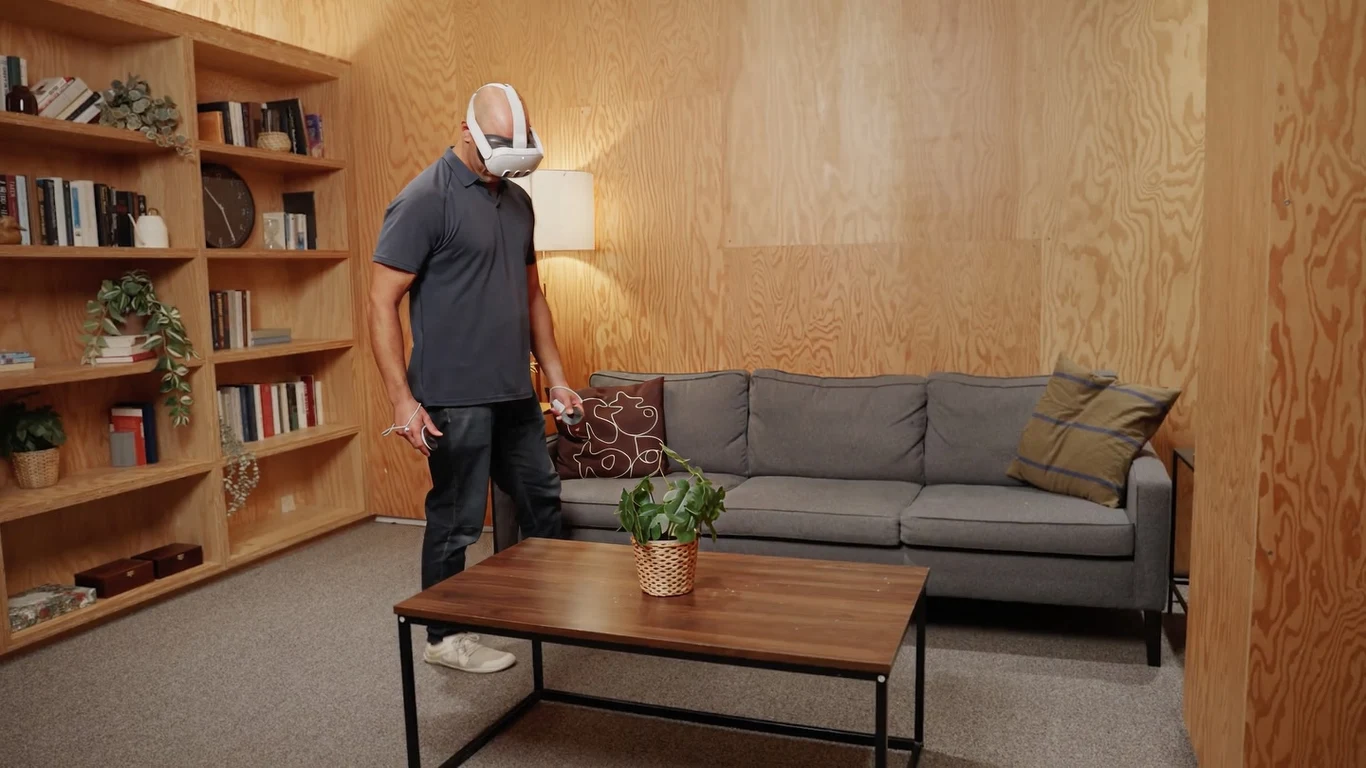 mężczyzna w goglach wirtualnej rzeczywistości stoi przy szarej sofie w minimalistycznym salonie z drewnianymi ścianami, na drewnianym stole stoi zielona roślina w wiklinowej doniczce, w tle widoczny regał z książkami i lampka stojąca