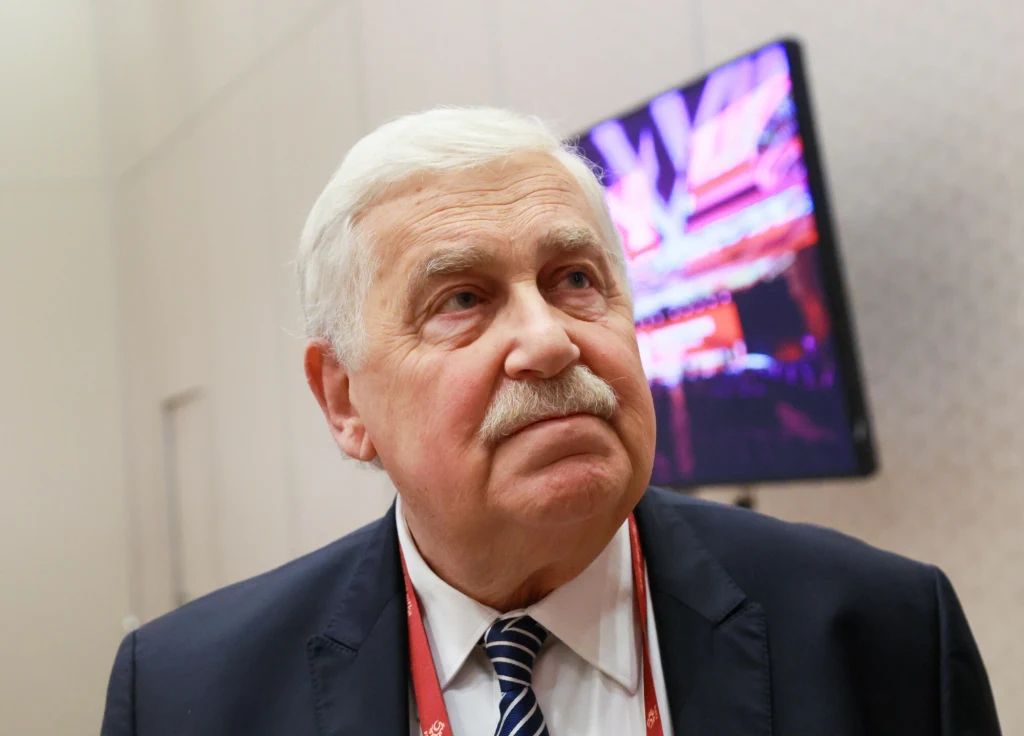 Jerzy Engel Starszy mężczyzna z siwymi włosami i wąsami w garniturze i krawacie stoi na tle rozmytego ekranu telewizora emitującego kolorowe światła.