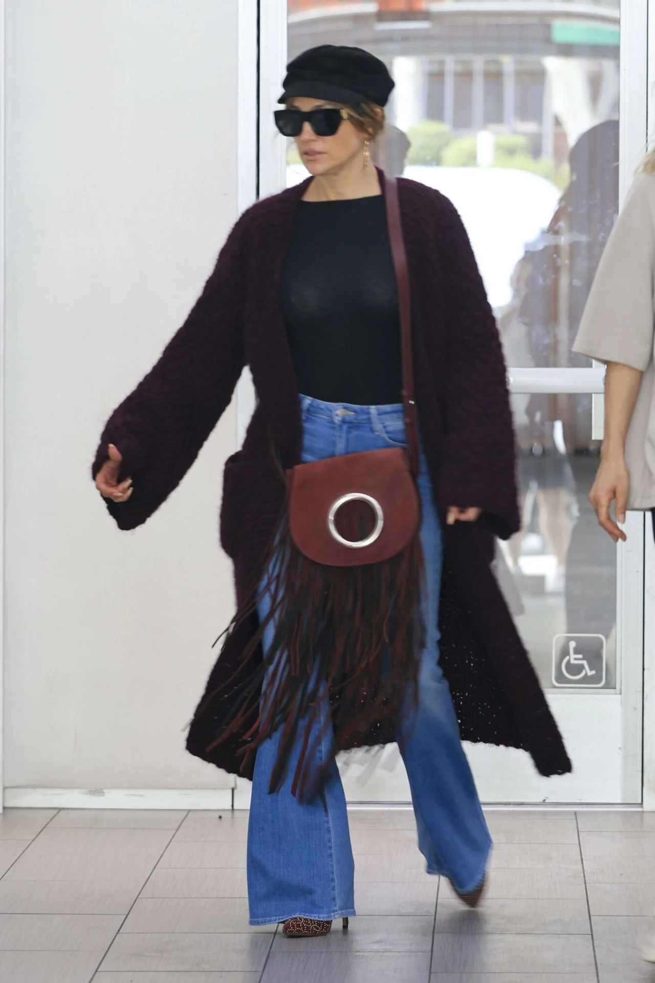 Jennifer Lopez w dzwonach i modnej stylizacji na jesień Kobieta ubrana w czarny kapelusz, duże okulary przeciwsłoneczne, bordowy płaszcz, czarną bluzkę i niebieskie dzwony, z torebką na pasku z frędzlami stoi przy wejściu do budynku.