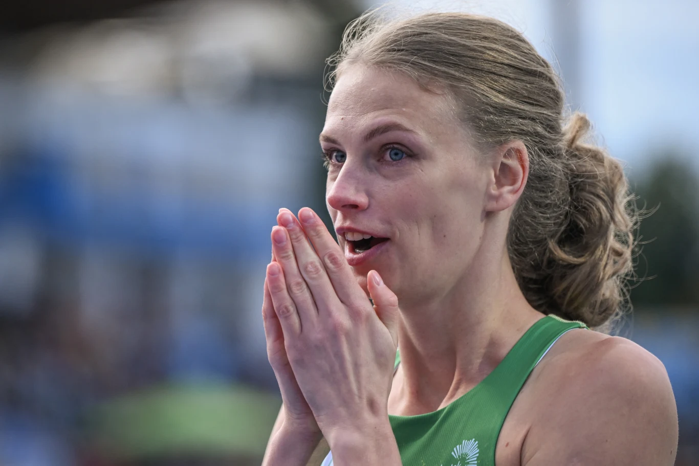 Kobieta w sportowym stroju zielonego koloru z zaskoczonym wyrazem twarzy, przykłada dłonie do ust w geście emocji, tło rozmyte, sugerujące stadion sportowy.
