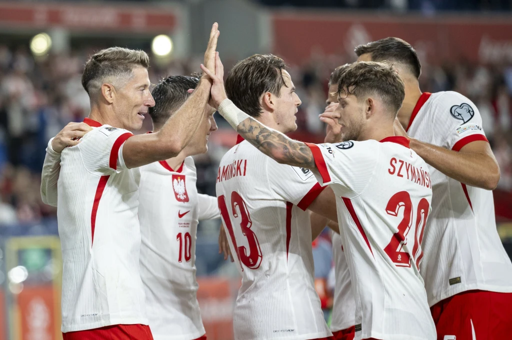 Reprezentacja Polski Grupa polskich piłkarzy w biało-czerwonych strojach świętuje zdobycie gola na stadionie, zawodnicy przybijają sobie piątki i obejmują się, w tle rozmyta widownia.