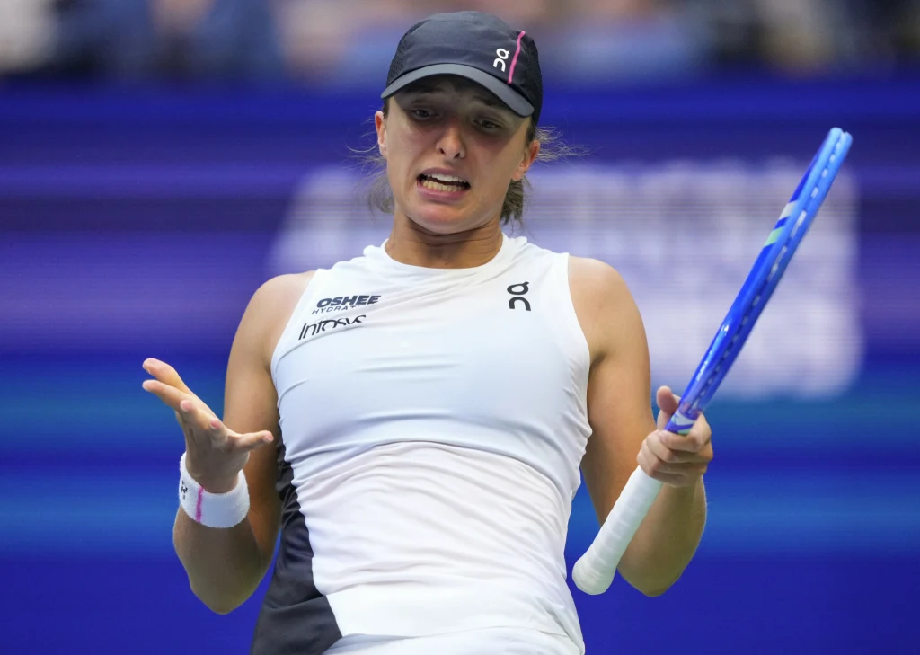 Tenisistka w białym stroju z czarnym daszkiem na głowie trzyma niebieską rakietę do tenisa, wykonując dynamiczny gest podczas gry, na tle rozmazanej niebieskiej ściany kortu.