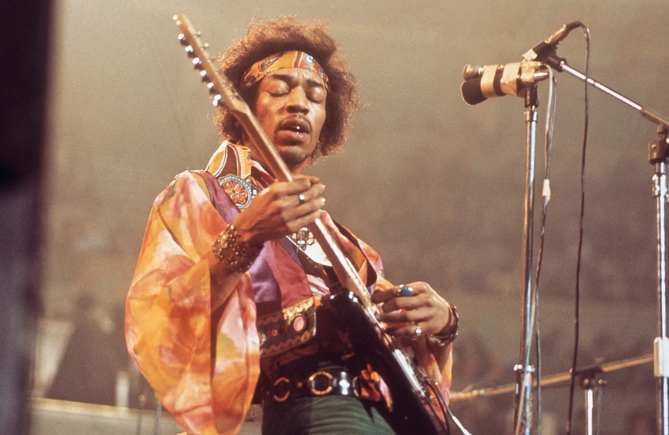 Jimi Hendrix zmarł w wieku 27 lat Legendarny gitarzysta w kolorowej koszuli i opasce na głowie podczas intensywnego występu scenicznego gra na gitarze elektrycznej obok mikrofonu, w tle widoczne światła sceniczne.