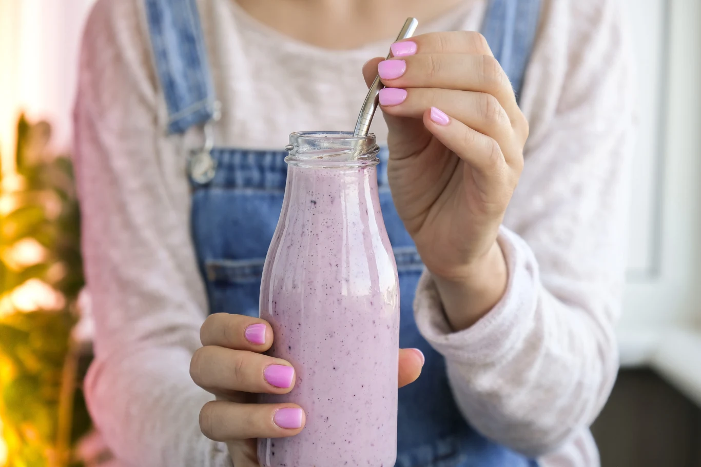 Kobieta w jasnej bluzce i ogrodniczkach trzyma szklaną butelkę z fioletowym smoothie oraz metalową słomkę, jej paznokcie pomalowane są na różowo.