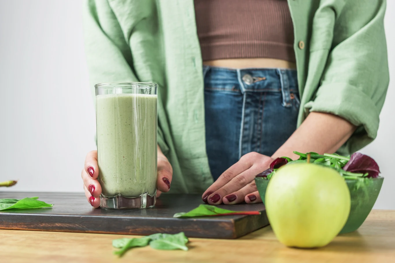 Szklanka zielonego smoothie trzymana przez osobę w zielonej koszuli, obok miska z sałatą i zielone jabłko ustawione na drewnianym blacie.
