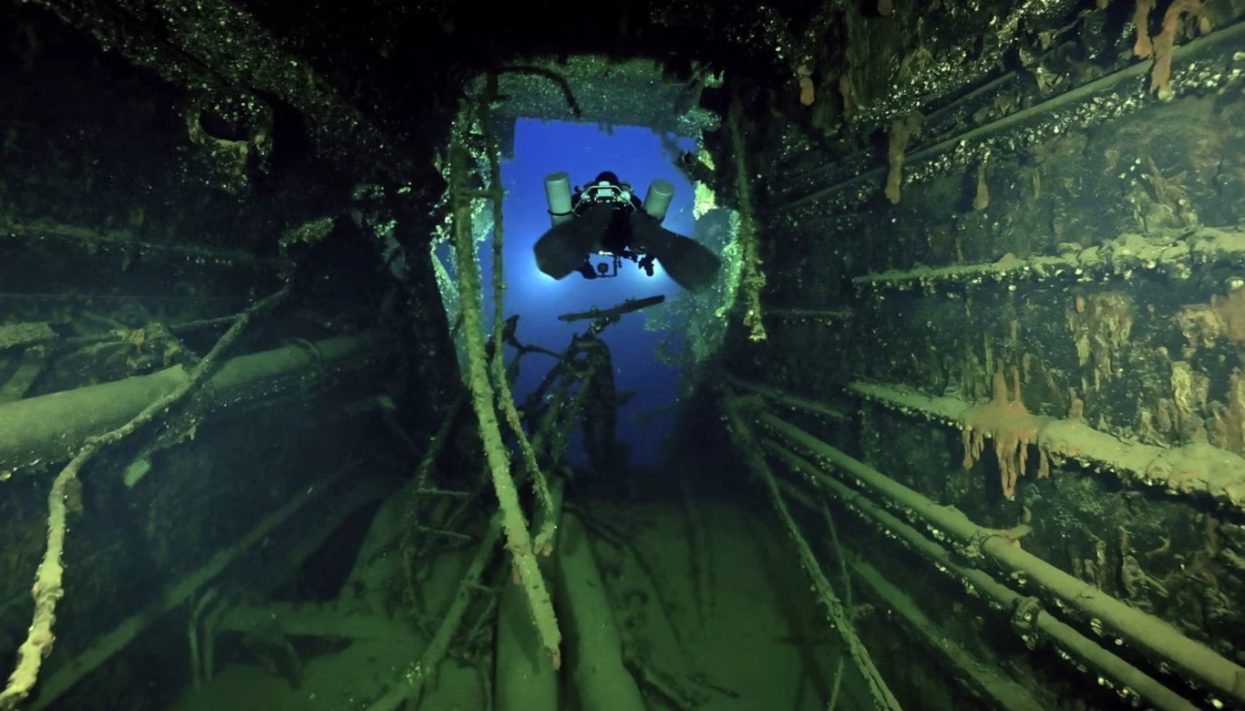 Nurkowie oświetlają wnętrze wraku HMHS Britannic po raz pierwszy od zatonięcia na Morzu Egejskim podczas I wojny światowej Nurek eksplorujący wnętrze zatopionego wraku, otoczony przez metalowe elementy konstrukcyjne porośnięte glonami i korozją, w tle widoczne niebieskie światło sugerujące otwarte wyjście lub większą przestrzeń.