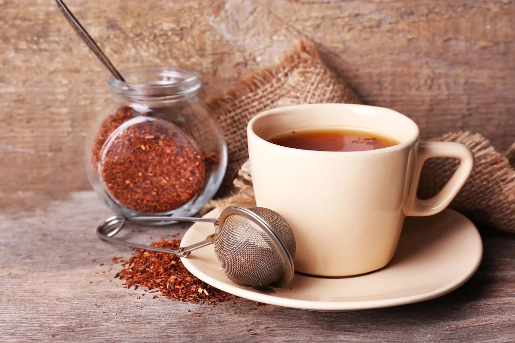 Herbata rooibos działa łagodniej niż kawa, przy okazji wspiera też naszą odporność i zdrowie Kubek z zaparzoną herbatą na spodku, obok szklany słoik z suszem herbacianym oraz metalowe sitko, całość na drewnianym stole z jutowym kawałkiem materiału w tle.