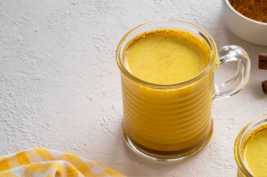 "Złote Mleko" świetnie rozgrzewa i obłędnie smakuje Przezroczysty kubek wypełniony żółtym napojem, którym jest złote mleko na bazie mleka i kurkumy, umieszczony na jasnej powierzchni obok żółtej ściereczki w kratkę; w tle widoczna miseczka z przyprawą oraz kawałek laski cynamonu.