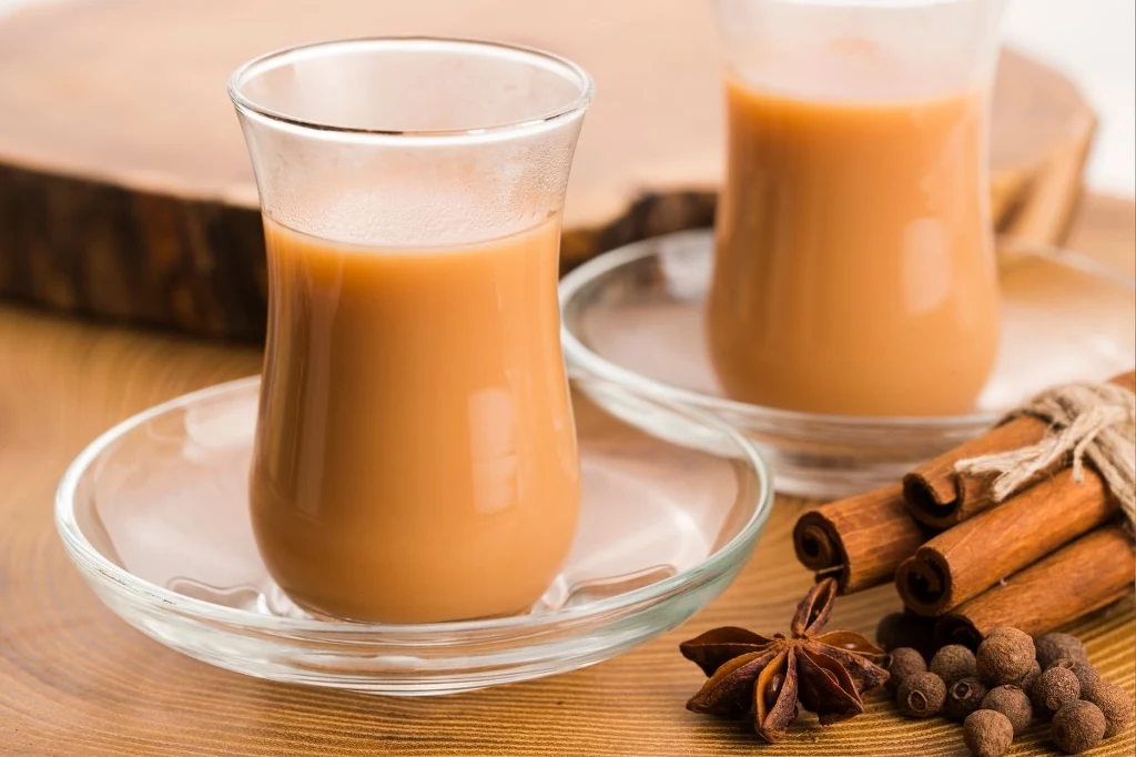 Masala chai oczarowuje aromatem Dwa szklane filiżanki z herbatą chai ustawione na przezroczystych spodkach, na pierwszym planie aromatyczne przyprawy: laski cynamonu, anyż gwiazdkowy i ziarna ziela angielskiego, tło drewniane w naturalnych odcieniach.