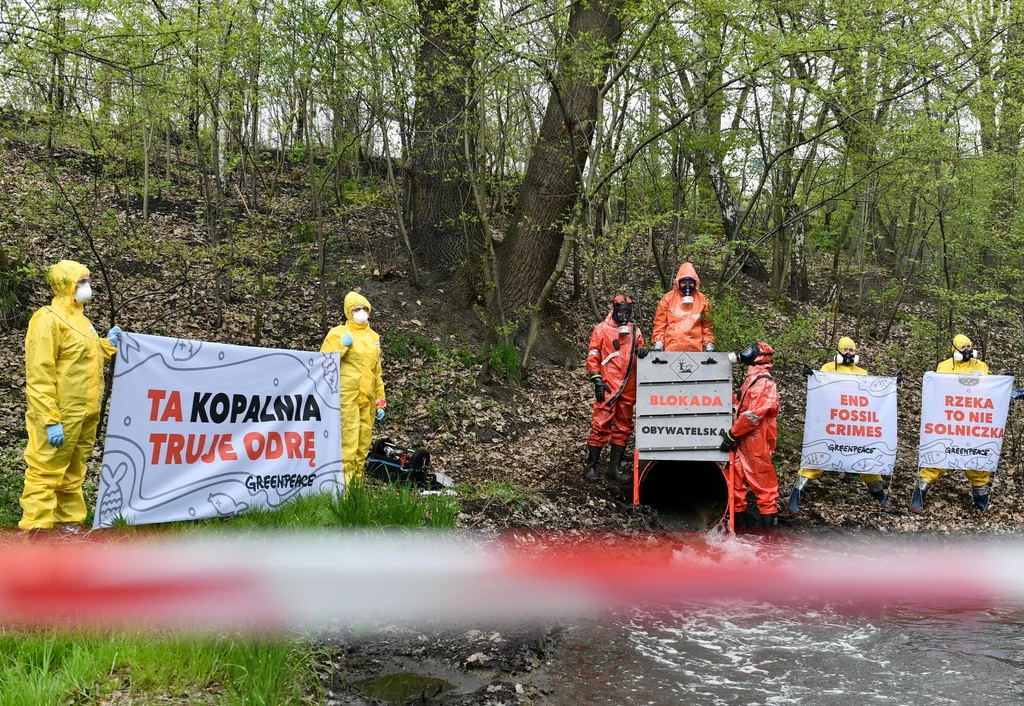 Grupa osób w kombinezonach ochronnych i maskach trzyma transparenty z hasłami ekologicznymi przy ujściu rury do rzeki w otoczeniu zieleni; na jednym z transparentów znajduje się napis 'TA KOPALNIA TRUJE ODRĘ', inne zawierają hasła przeciwko spalaniu pa...