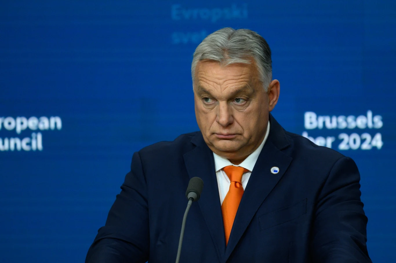 Premier Węgier Viktor Orban Mężczyzna w garniturze z pomarańczowym krawatem stoi przed niebieskim tłem z napisem Brussels oraz datą 2024, patrząc poważnie na wprost, mówiąc do mikrofonu.