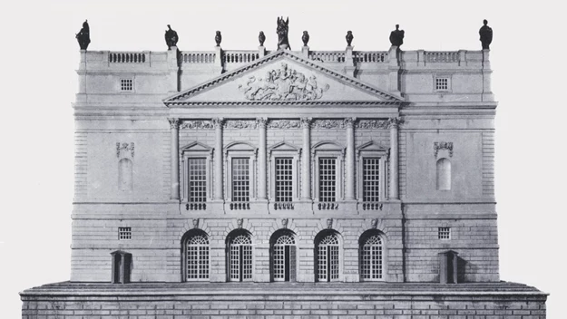 Monumentalna fasada klasycystycznego budynku ozdobiona kolumnami, licznymi rzeźbami oraz detalami architektonicznymi, z symetrycznie rozmieszczonymi oknami i wysokim cokołem u podstawy.