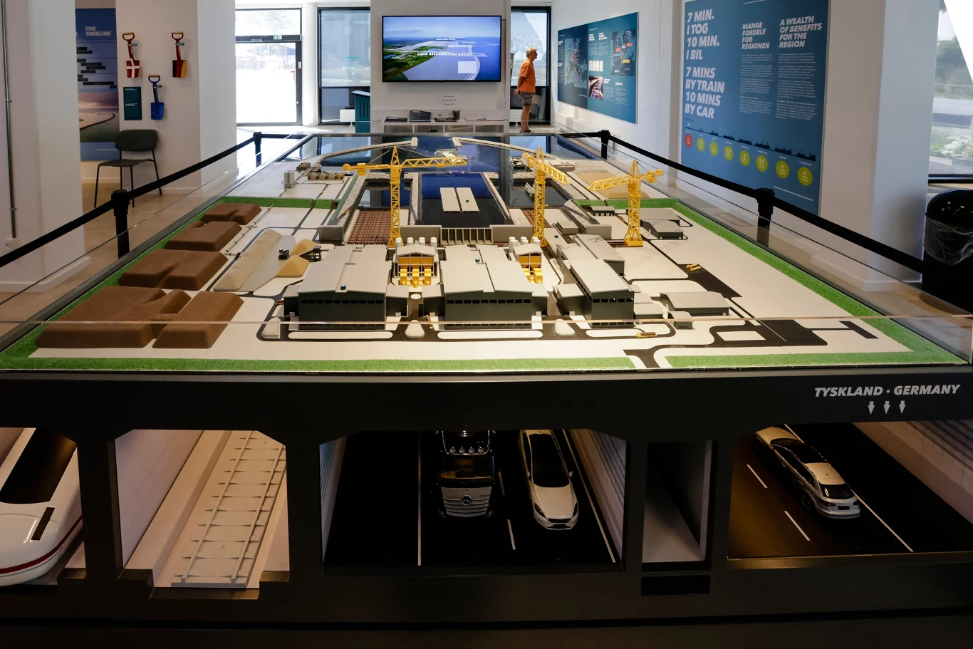 W tunelu będą dwie dwupasmowe jezdnie i dwutorowa linia kolejowa Makieta nowoczesnego centrum logistycznego z rozmieszczonymi żółtymi dźwigami, modelami hal magazynowych i pojazdami pod powierzchnią, znajdująca się w jasnym wnętrzu ekspozycyjnym.