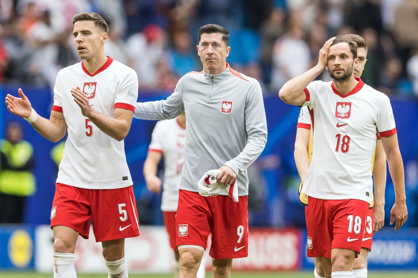 Bartosz Bereszyński (z prawej) Trzech piłkarzy reprezentacji Polski w białych koszulkach z czerwonymi spodenkami idzie razem po murawie boiska, jeden z nich opiera dłoń na ramieniu drugiego, a w tle widoczni są kibice.