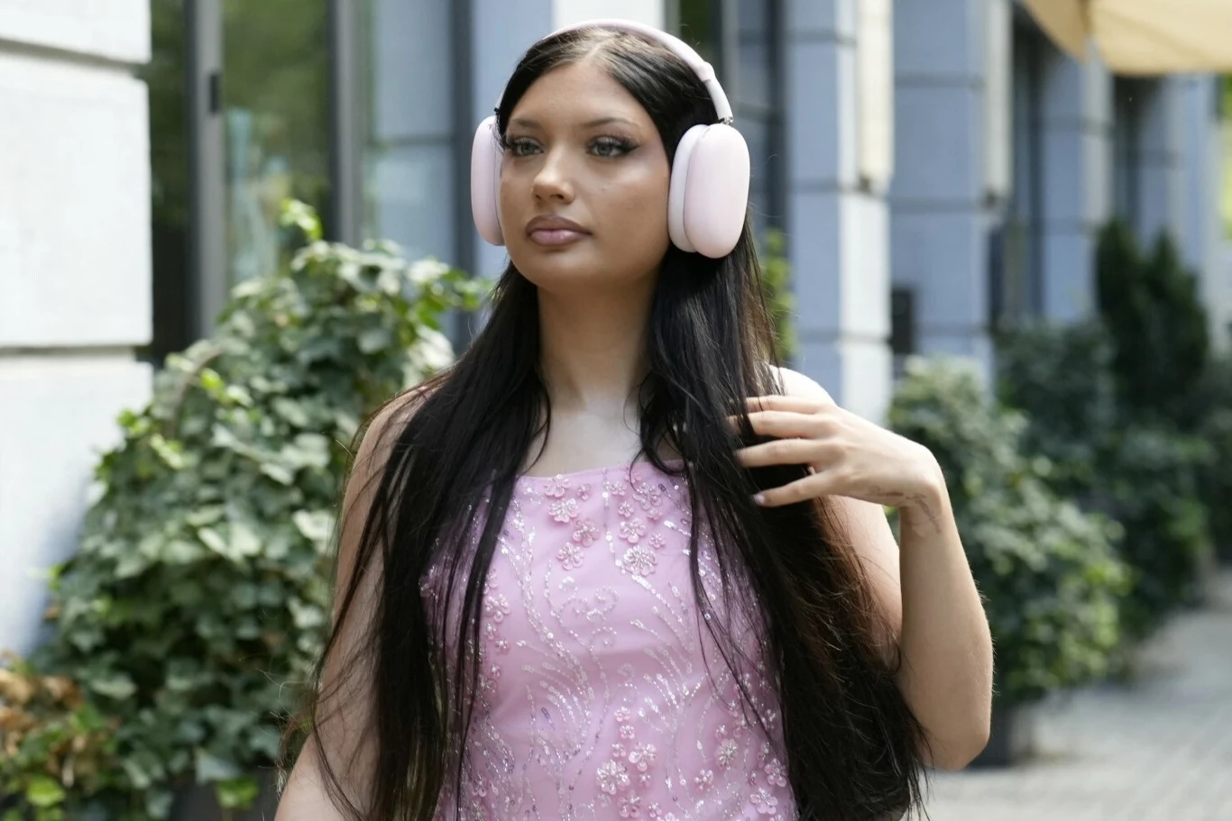 Viki Gabor Młoda kobieta z długimi ciemnymi włosami nosząca duże, jasnoróżowe słuchawki oraz różową sukienkę ozdobioną cekinami, spaceruje wzdłuż budynku z zielonymi krzewami w tle.