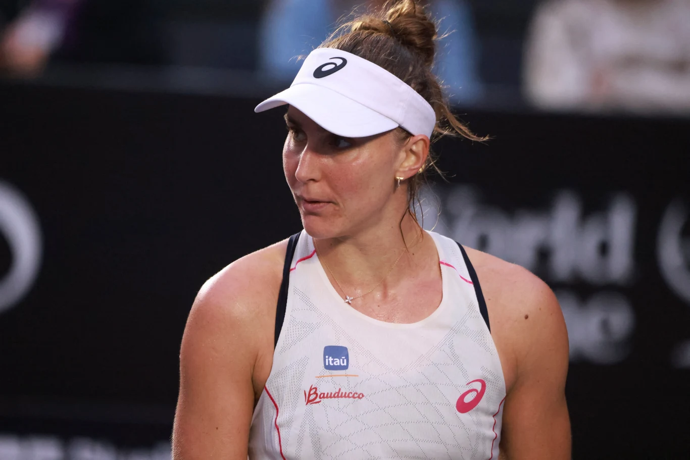 Kobieta w stroju sportowym i białej czapce z daszkiem patrzy w bok, na jej koszulce widoczne są loga sponsorów, tło stanowi rozmazana powierzchnia kortu oraz ciemne banery reklamowe.