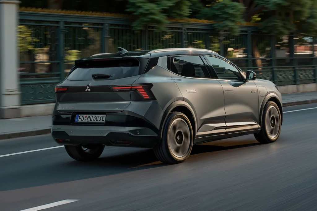 Mitsubishi Eclipse Cross nie jest autorską konstrukcją japońskiej marki. Bazą do jego stworzenia było Renault Scenic. Nowoczesny, szary samochód osobowy marki Mitsubishi porusza się po miejskiej ulicy, otoczony przez zieleń i metalowe ogrodzenie.